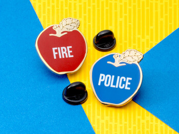 Red apple lapel pin with FIRE text and blue apple lapel pin with POLICE text, both featuring gold trim and black rubber backings.