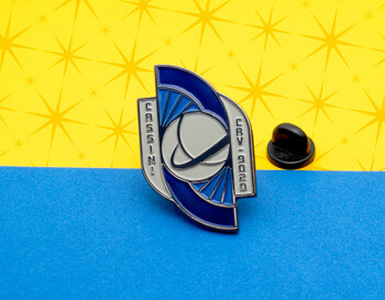 Blue and silver Cassini CRV 9020 enamel lapel pin with a planetary orbit design and black rubber clutch on a yellow and blue background.