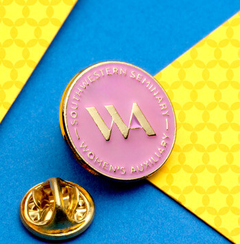 Circular pink and gold lapel pin for Southwestern Seminary Women's Auxiliary featuring a WA monogram on a blue and yellow patterned background.