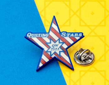 Star-shaped lapel pin with red and white diagonal stripes and blue border featuring Quilting Stars QW 2023 text.