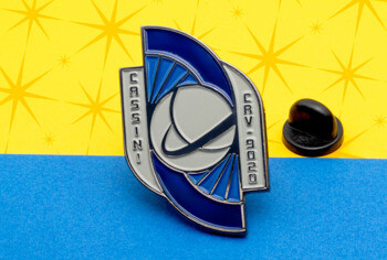 Blue and silver Cassini CRV 9020 enamel lapel pin with a planetary orbit design and black rubber clutch on a yellow and blue background.