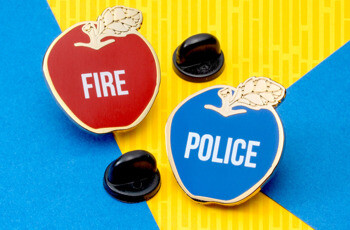 Red apple lapel pin with FIRE text and blue apple lapel pin with POLICE text, both featuring gold trim and black rubber backings.