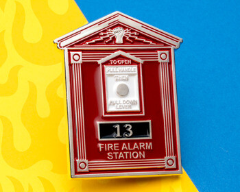 Red and silver lapel pin shaped like a vintage Fire Alarm Station box with number 13 on a yellow and blue background.