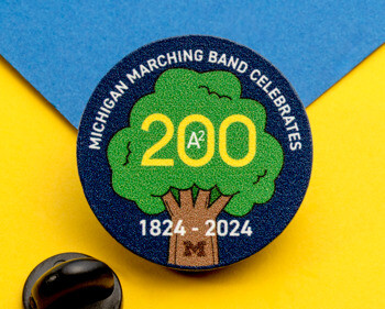 Circular lapel pin with Michigan Marching Band 200 design featuring a green tree and years 1824-2024 on a blue background.