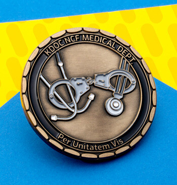 Circular challenge coin for KDOC NCF Medical Dept featuring a stethoscope and handcuffs design on an antiqued bronze finish.