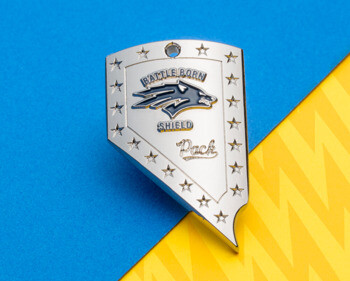Shield-shaped lapel pin with Battle Born Shield Pack text, a wolf logo, and star border on a blue and yellow background.