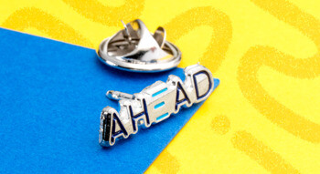 Silver die cast lapel pin featuring the word AHEAD with blue enameled accents on a yellow and blue background.