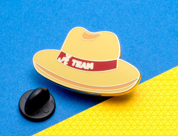 Yellow fedora hat lapel pin with a red band featuring the Alabama script A logo and the word TEAM, shown with a black rubber clutch on a blue and yellow backgro